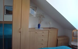 A room with a sloped ceiling, a wooden wardrobe with drawers, a small wicker side table, and a framed picture on the wall.