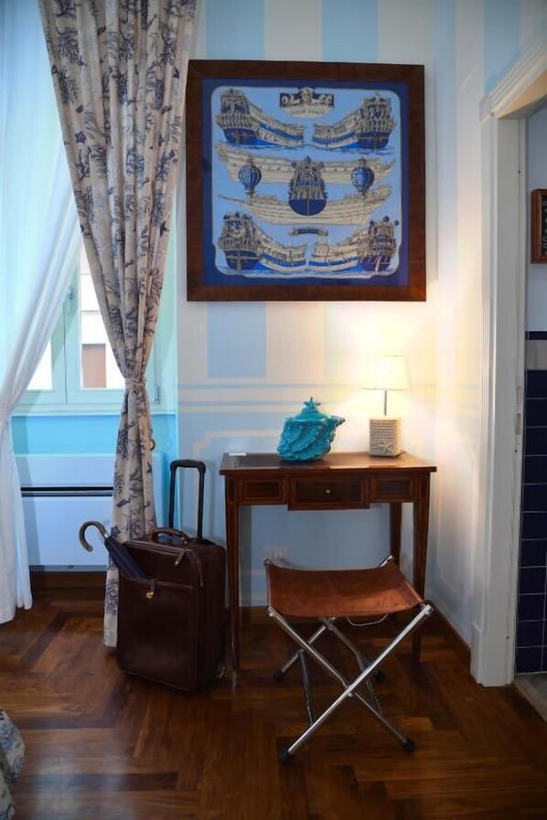 A room with a wooden floor, a desk with a lamp, a suitcase, a folding chair, and a framed picture on the wall.