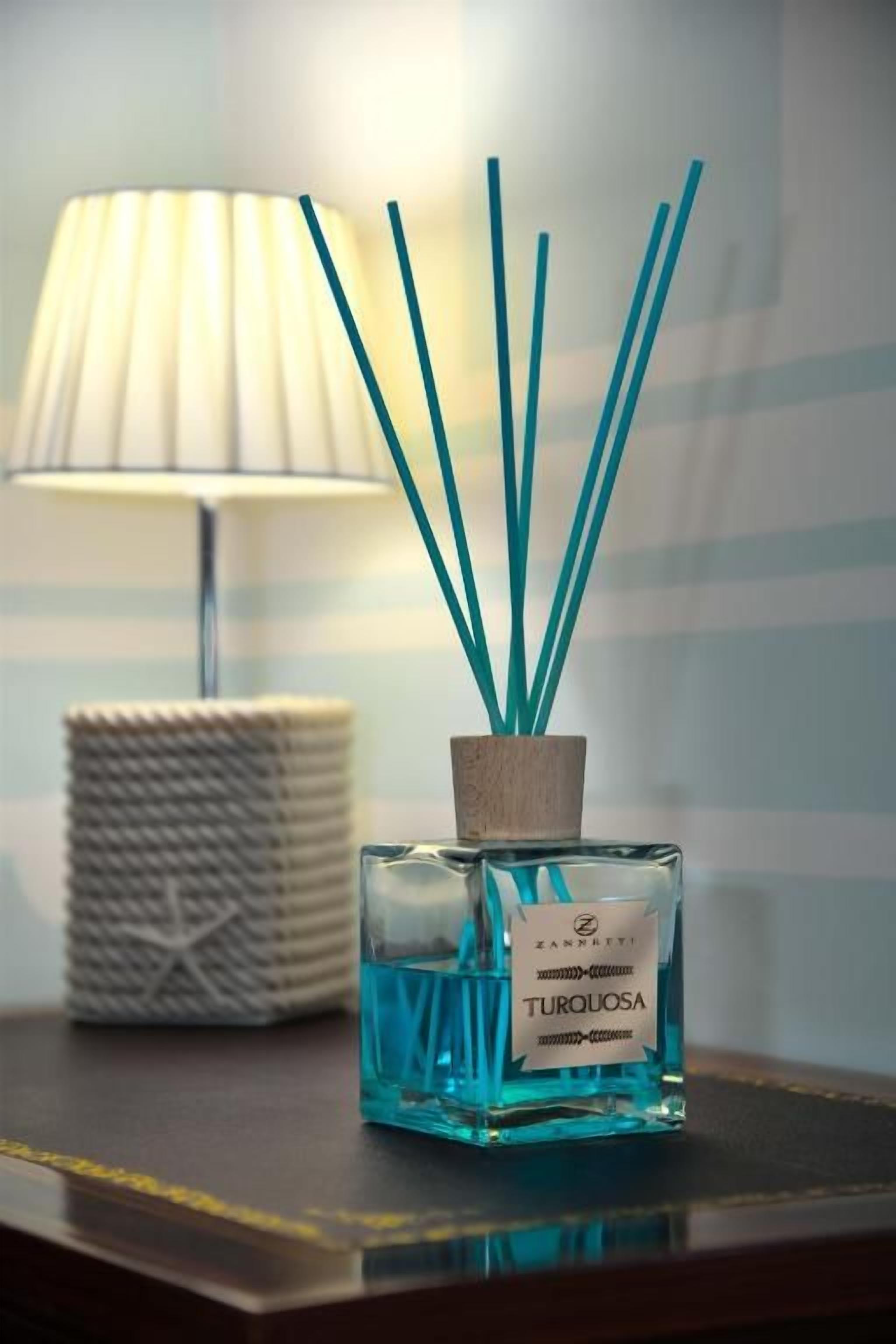 En flaske 'Turquoise' cologne med rørreder på et bord, en lampe og en flettet kurv i baggrunden.