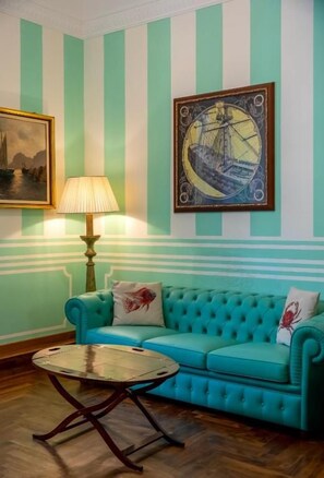 A room with a teal sofa, a round coffee table, a lamp, and two framed artworks on the wall. - Regal House Anzio Manetti 16 (Anzio)