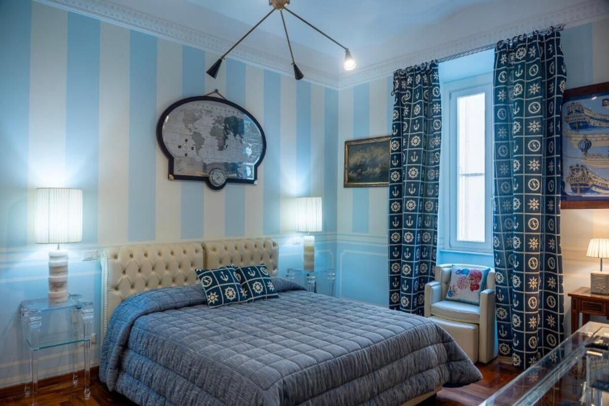 A bedroom with a tufted headboard, a large bed with a blue quilt, a wall clock, and striped wallpaper.