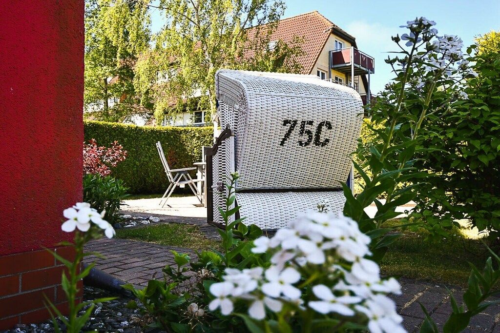 A white outdoor storage unit with the number '75C' on it, placed in a garden area with a red wall and greenery.