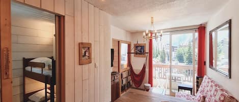 A room with a bunk bed, wooden walls, a dining table, and a view through a window.