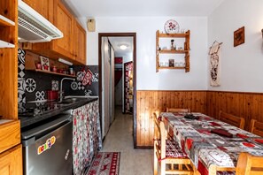 Cocina compacta con estufa, lavamanos y armarios de madera. Se ve un comedor con mesa y sillas.