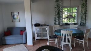 Ein Wohnzimmer mit einer Couch, einem Esstisch mit Stühlen, einem Schrank mit Glastüren und einem Fenster mit Vorhängen.