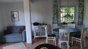 Ein Wohnzimmer mit einer Couch, einem Esstisch mit Stühlen, einem Schrank mit Glastüren und einem Fenster mit Vorhängen.
