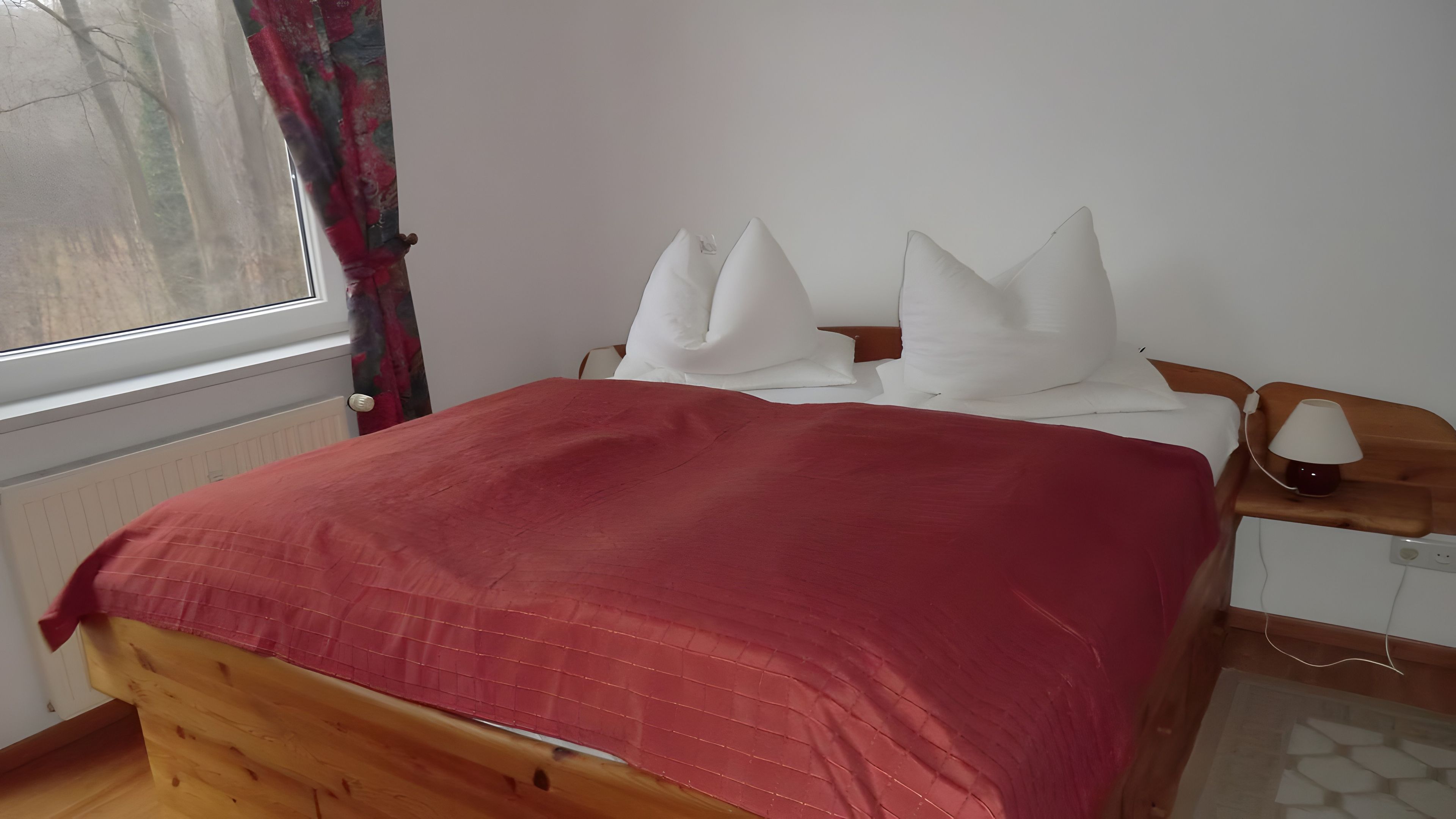 A bed with a red blanket, white pillows, and a bedside table with a lamp.