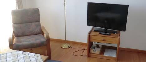 Een kamer met een bank, een televisie op een houten dressoir, een vloerlamp en een bed met een gestreept dekbed.