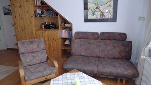 A living room with a wooden bookshelf, a patterned sofa, and a table with a grid-patterned tablecloth. - Apartment 'Koserow Seebad, Waldstraße 14' with Balcony and Wi-Fi (Koserow)