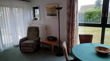 A room with a chair, a table with a basket, a floor lamp, a radiator, and a window with curtains.