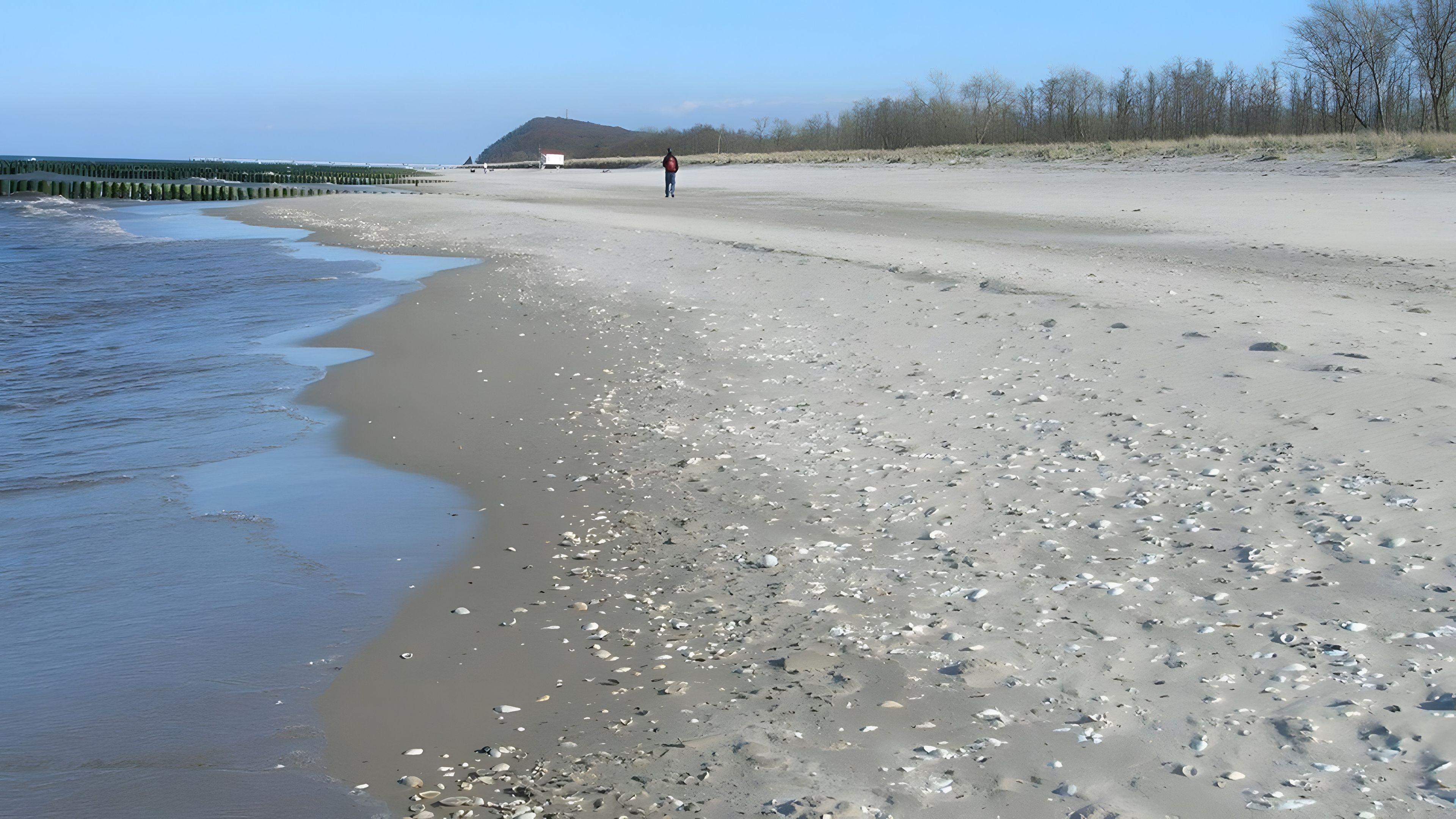 Une plage où l’on voit une personne qui se promène, des galets et une colline au loin.