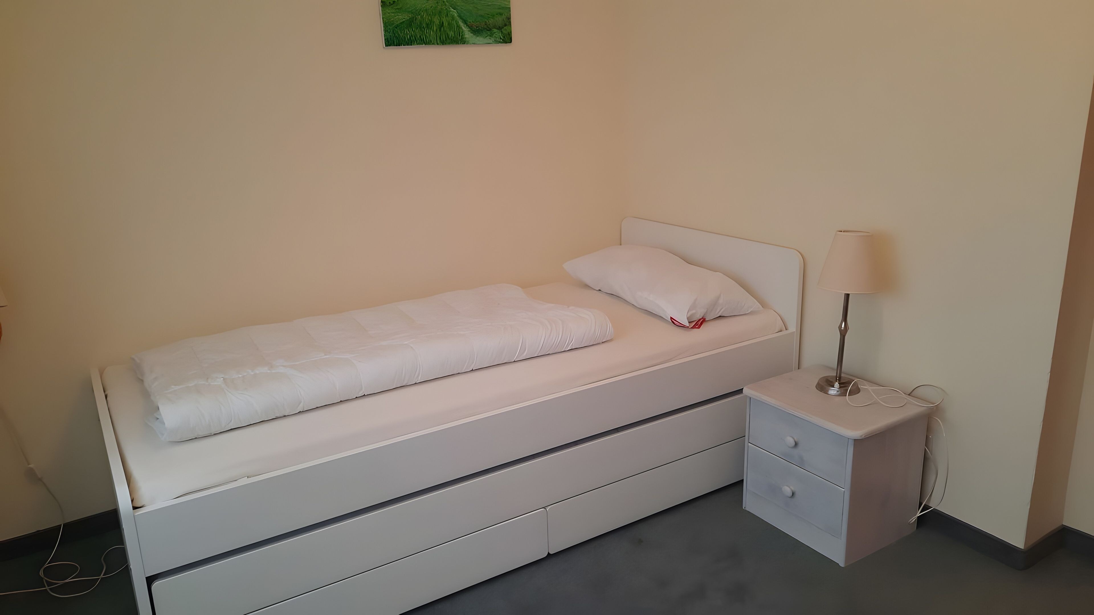 A single bed with a white mattress and pillows, a bedside table with a lamp, and a small drawer unit.