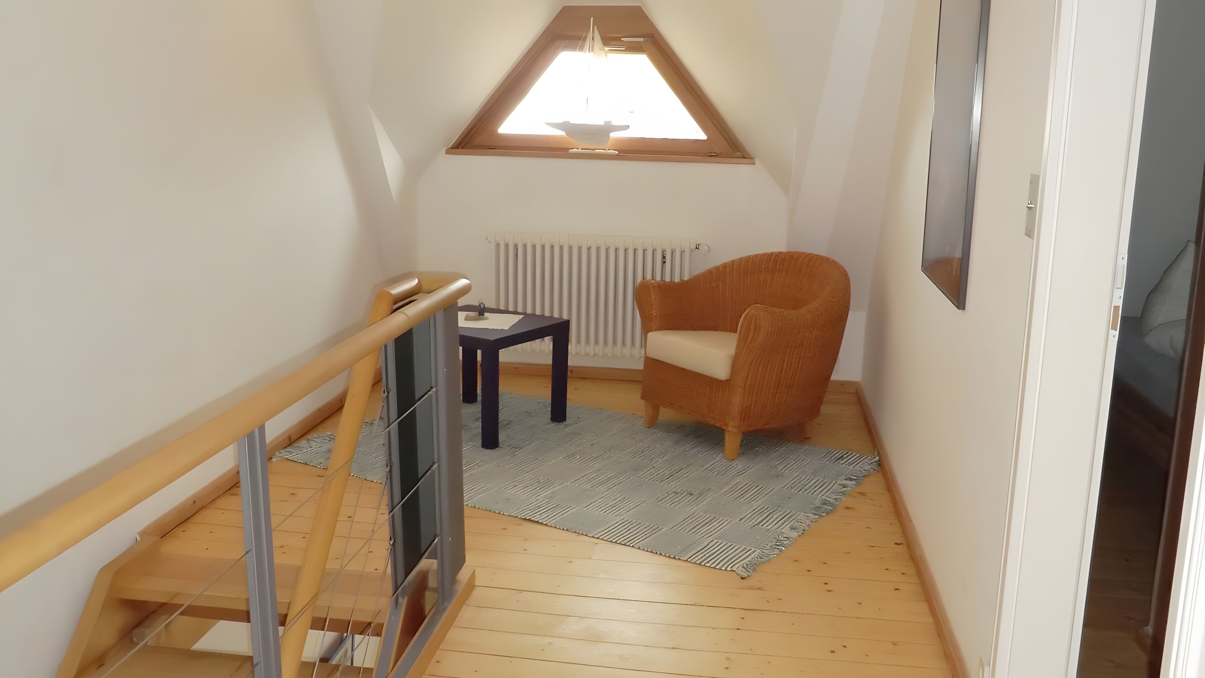 Un pequeño altillo bien iluminado con techo inclinado, una silla de madera, una mesita y un radiador.