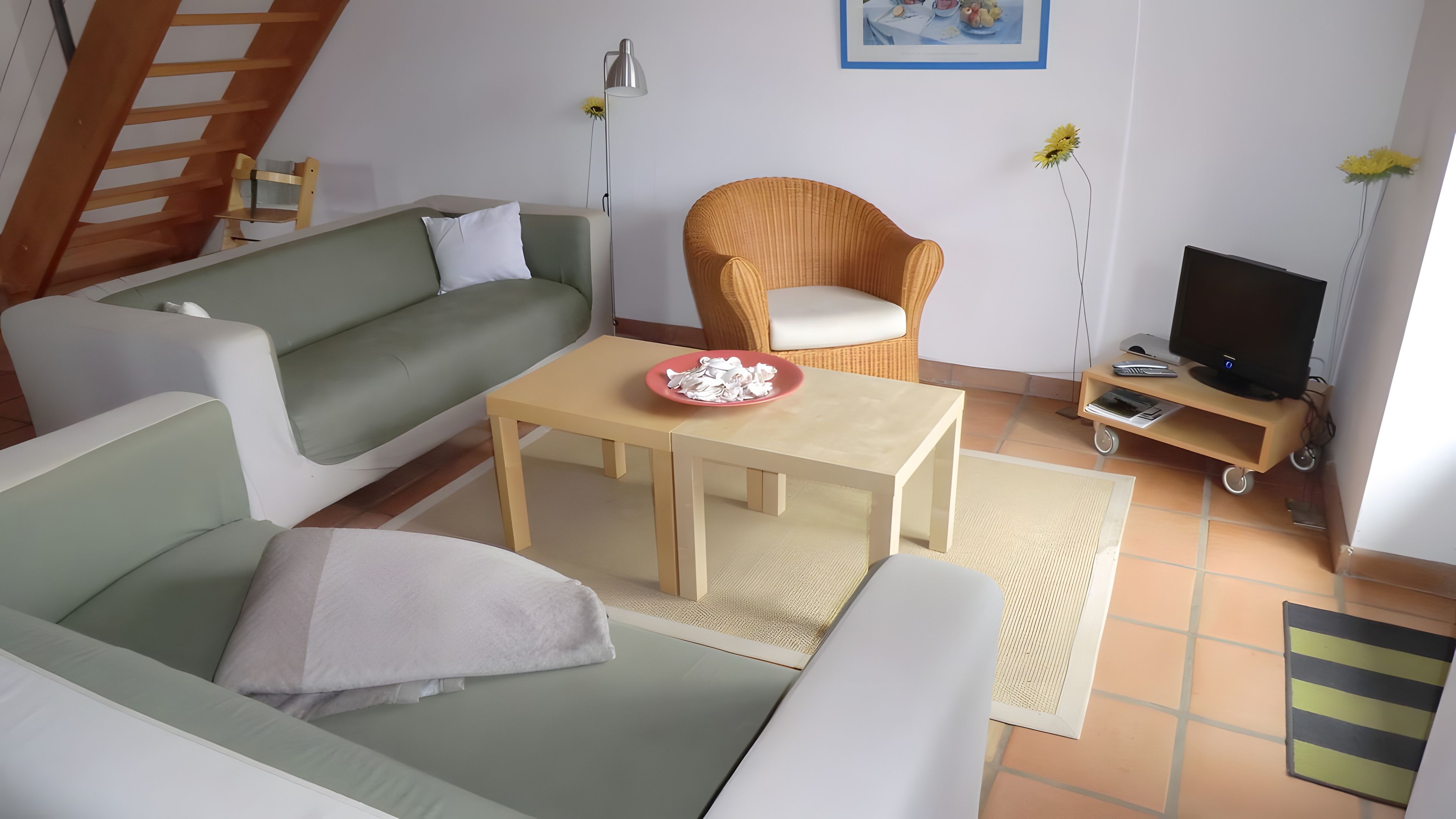 Un salon avec un canapé vert, une table basse en bois, un fauteuil en osier, un téléviseur posé sur un meuble et un tableau accroché au mur.