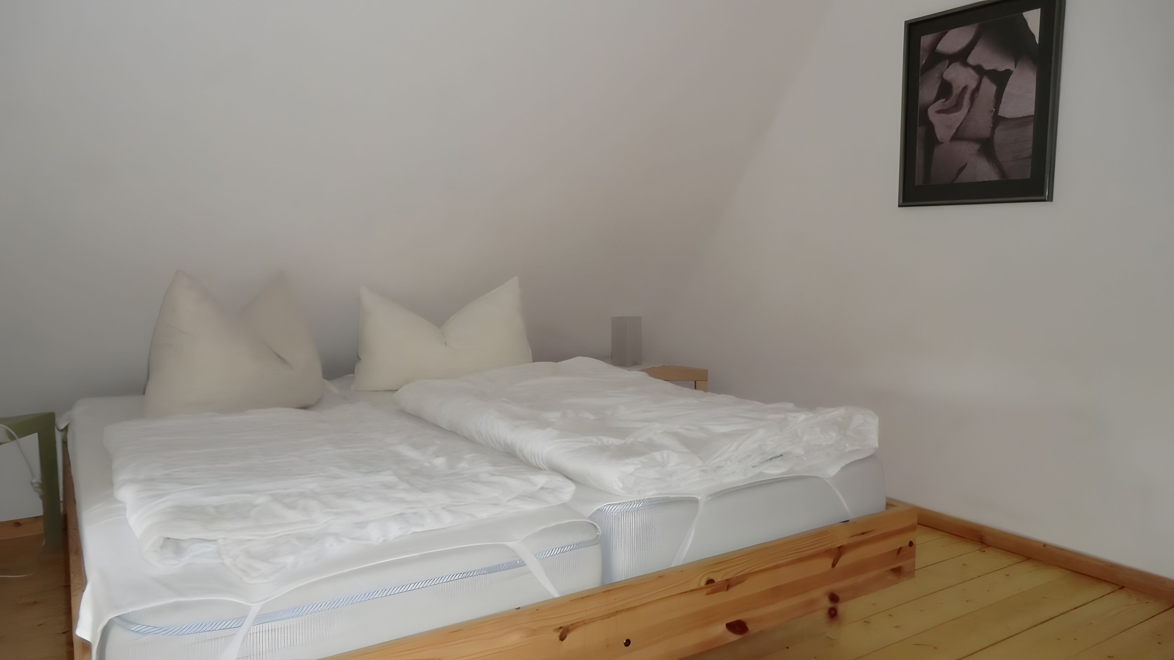 Una cama doble con ropa blanca en una habitación con suelo de madera y un cuadro enmarcado en la pared.