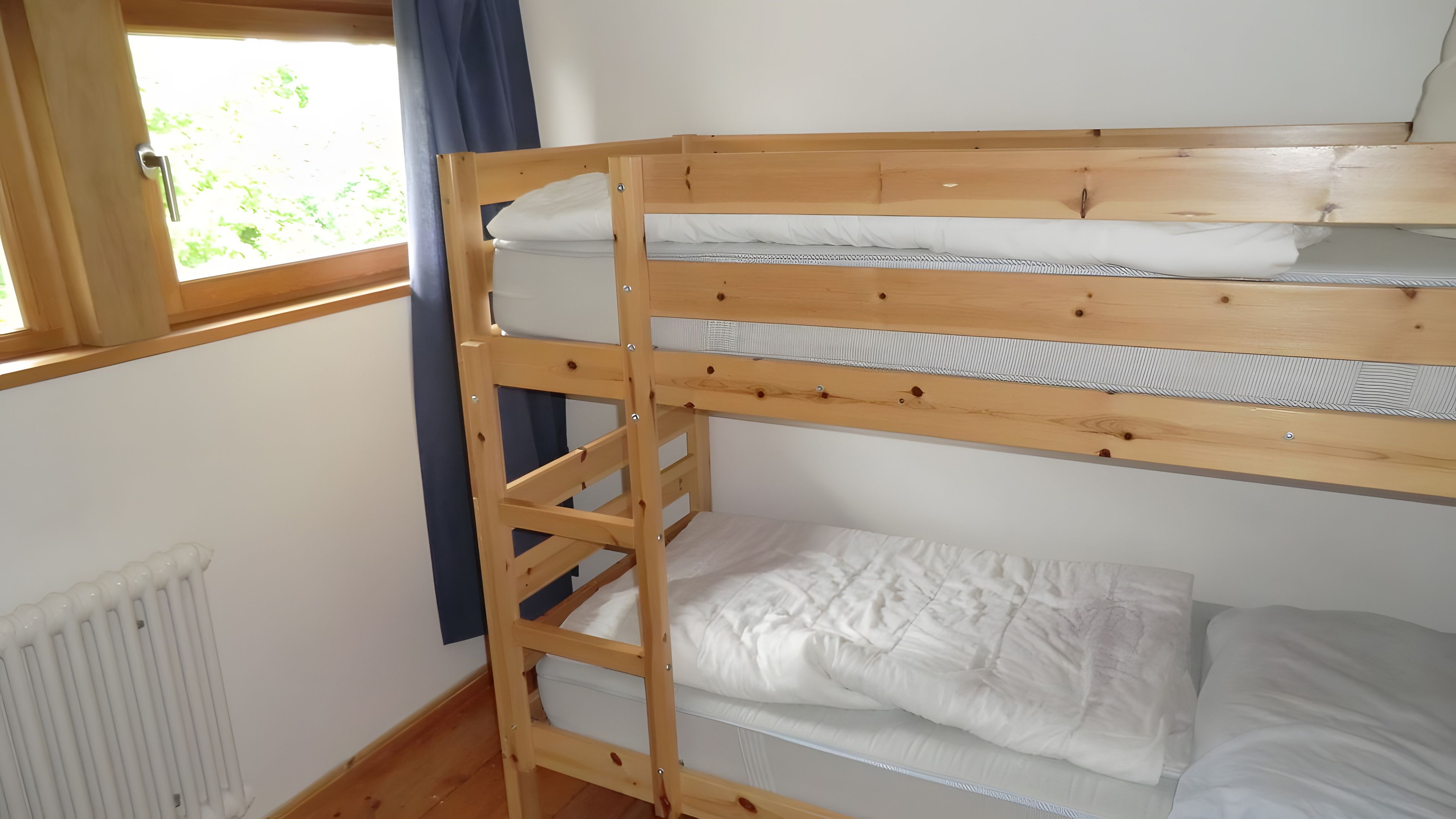 Una cama nido con escalera, ropa de cama blanca, una ventana con cortinas y un radiador.