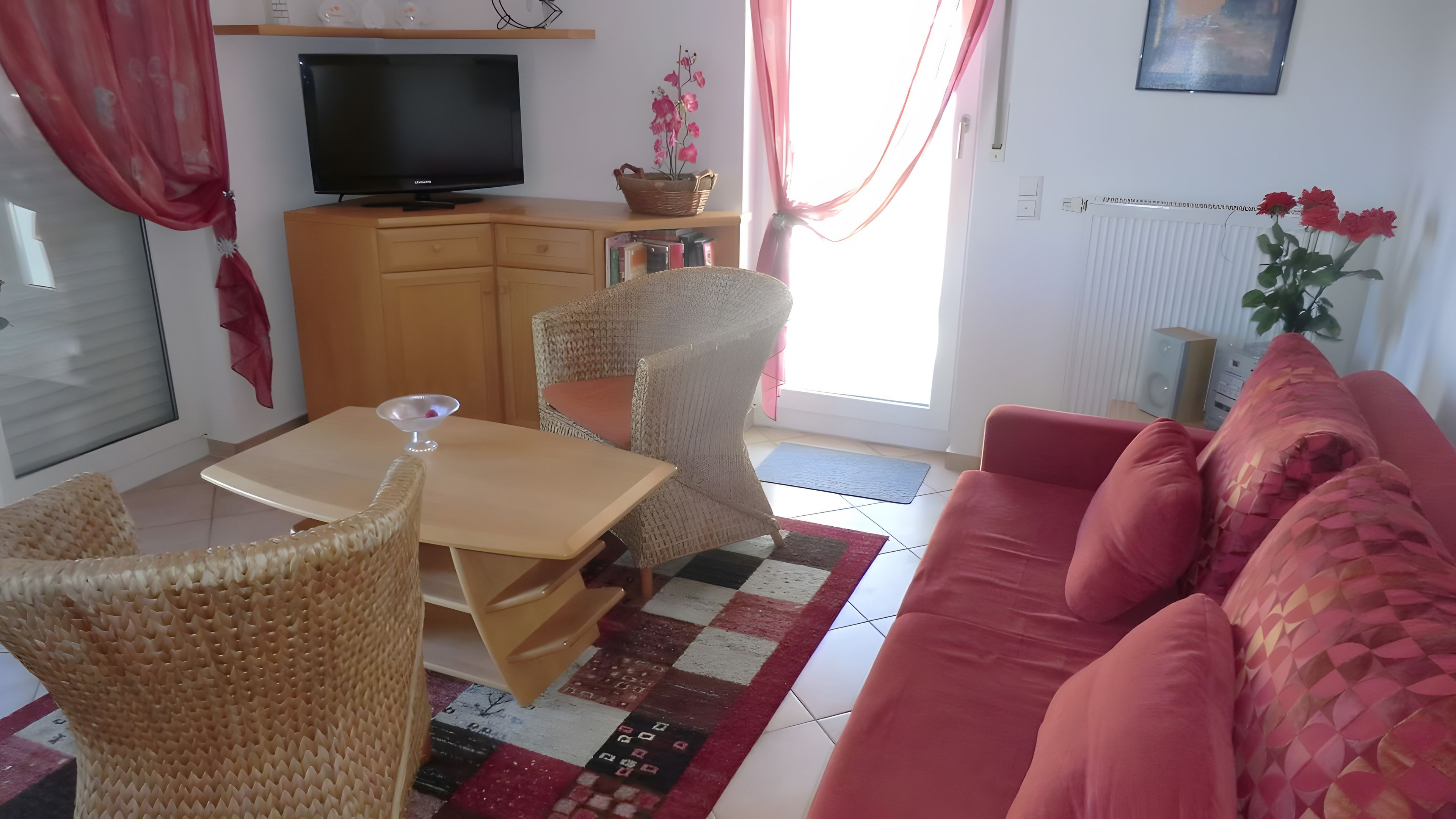 Ein Wohnzimmer mit einer Couch, einem Fernseher, Korbstühlen, einem Tisch und einem roten Teppich.