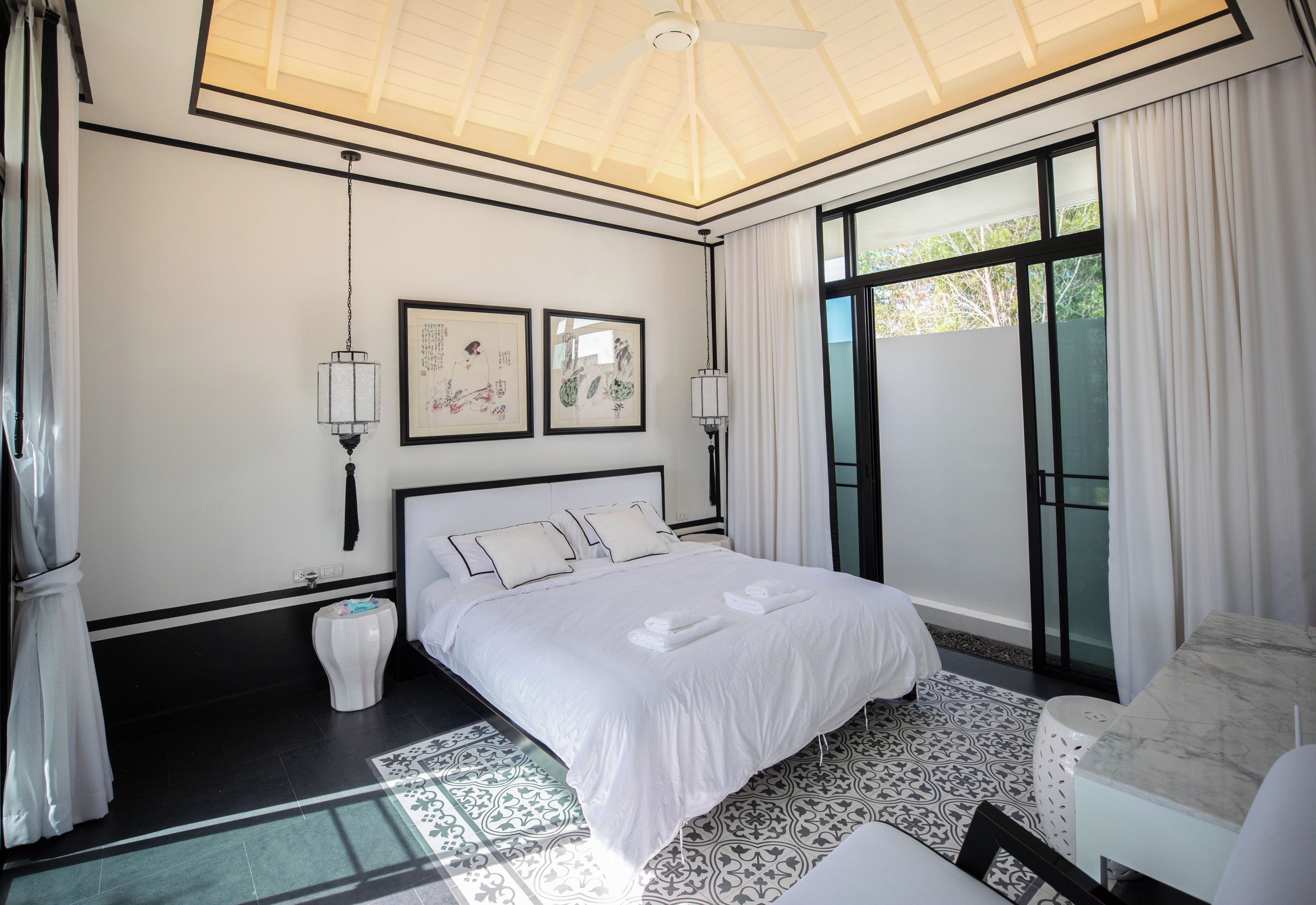 Una habitación de hotel con una cama grande, dos lámparas fijadas a la pared, un ventilador de techo y un ventanal con cortinas.