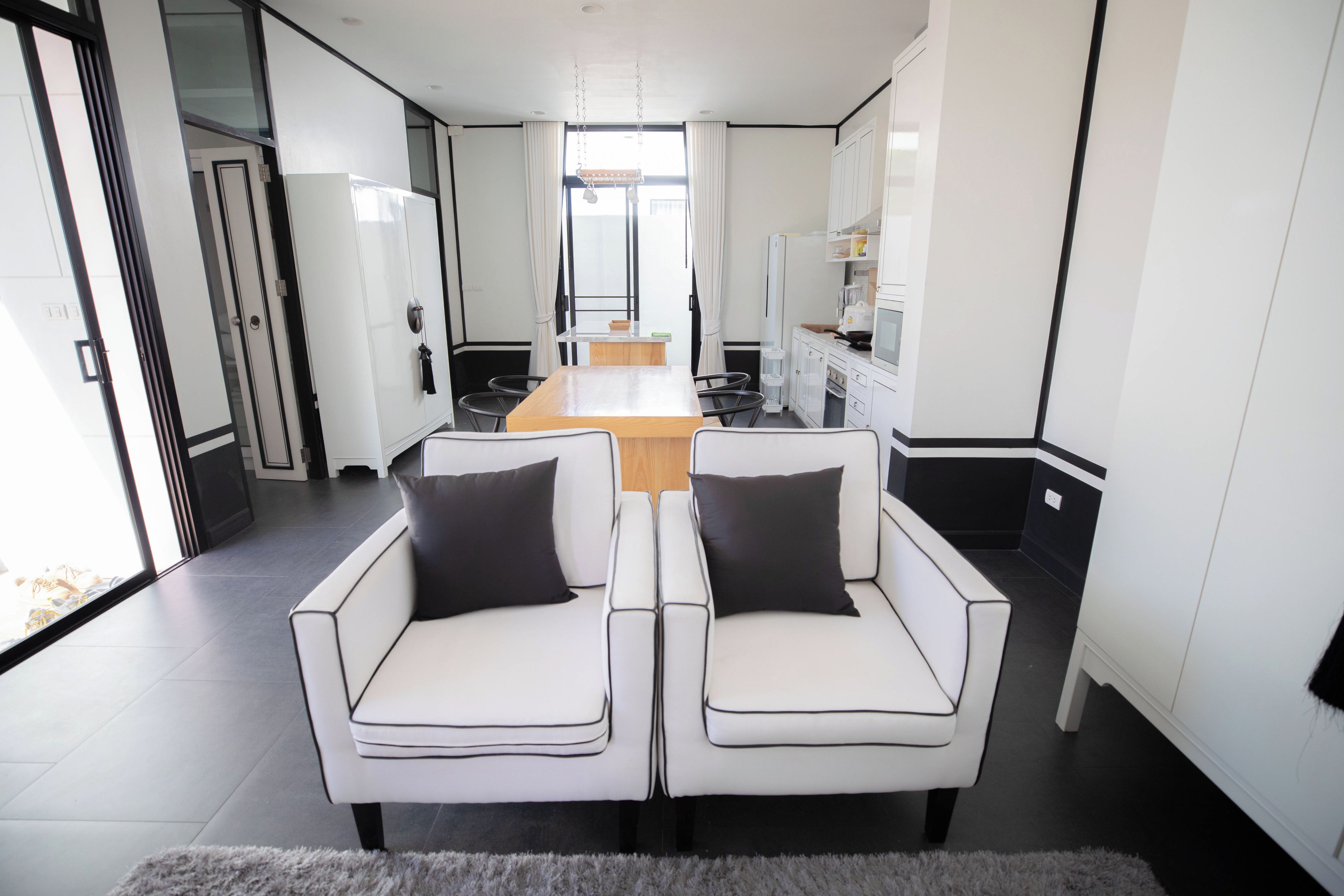 Een moderne keuken met witte kasten, een eethoek met een houten tafel en twee witte stoelen met zwarte kussens, en een groot raam dat zorgt voor daglicht.