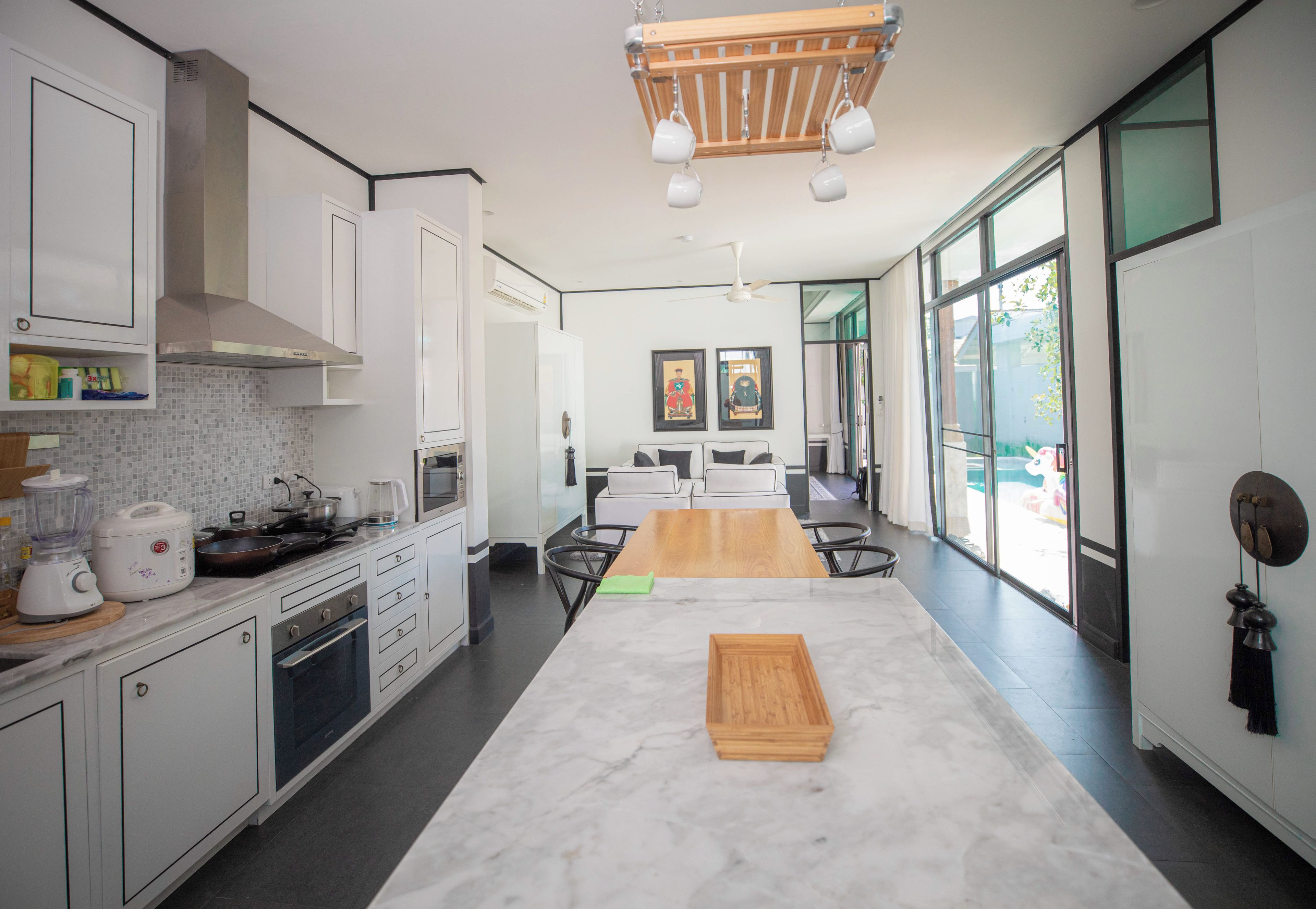 A modern kitchen with white cabinets, a wooden dining table, and a view through large windows.