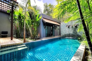 Un área de piscina moderna al aire libre con un muro de piedra, un espacio de estar cubierto y exuberante vegetación tropical.