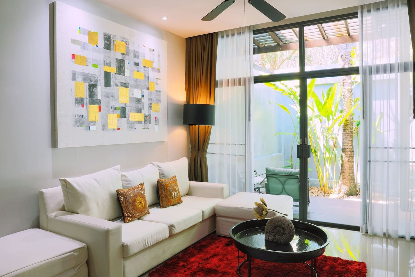 A modern living room with a white sofa, a red rug, a black coffee table with a vase, and a large window with curtains.