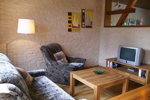 A cozy living room with a brick wall, a patterned armchair, a wooden coffee table, a television, and a lamp. - Holiday apartment with 1 bedroom (Ostseebad Prerow)
