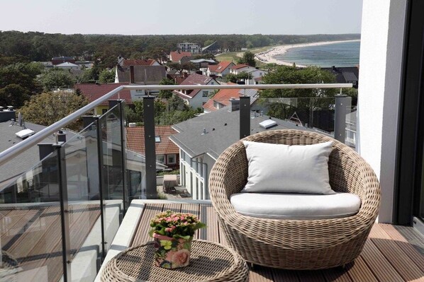 Un balcon avec un fauteuil en osier et une petite table, donnant sur une plage et un quartier résidentiel.