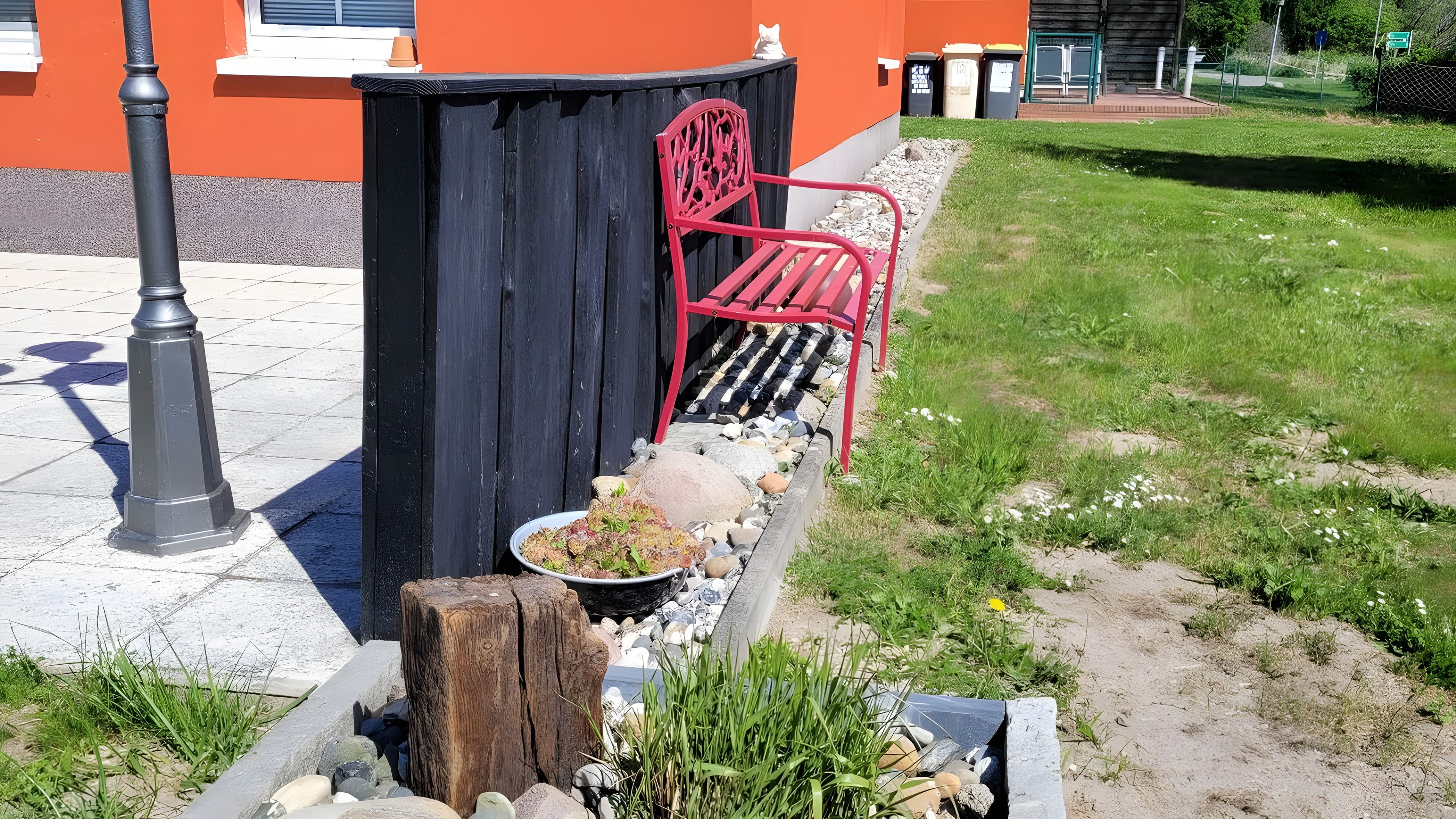 Un'area giardino con un cestino nero, una sedia rossa e un palo di legno.