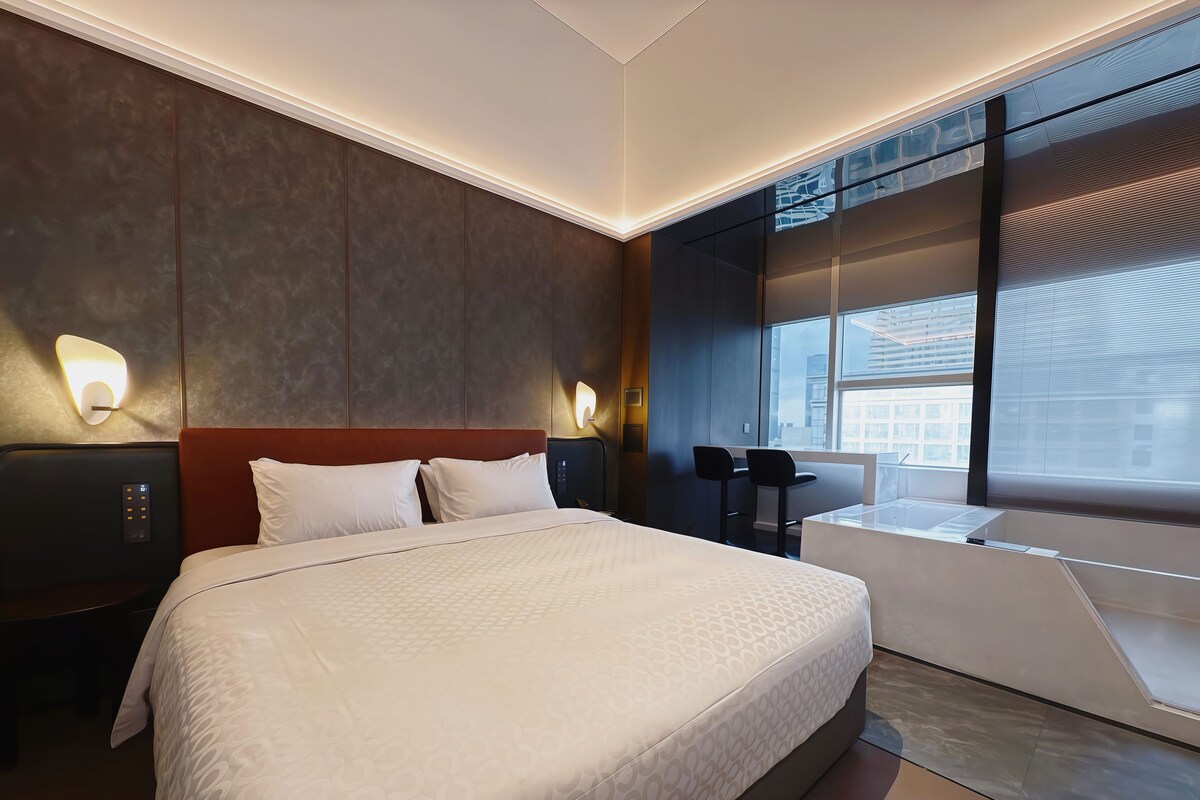 A modern hotel room with a large bed, bedside tables, a desk, and a window with a view of the city.