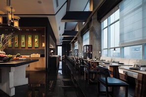 A modern restaurant with a long counter, wooden chairs, and a view of the city through large windows.