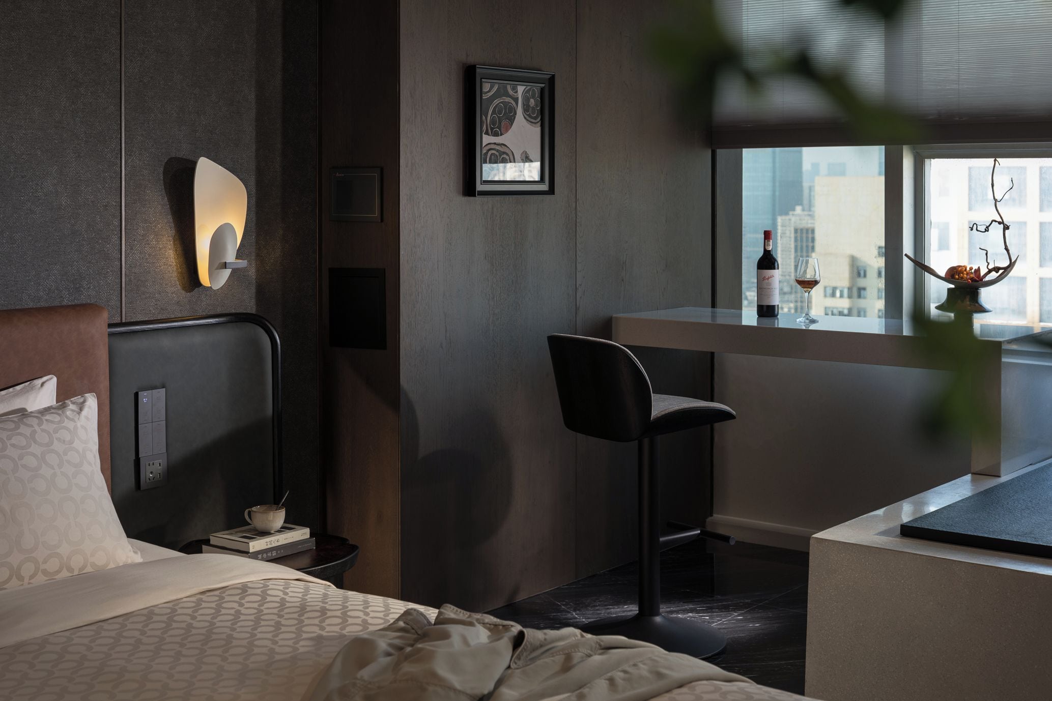 A modern hotel room with a bed, a desk, a chair, a window with a city view, and a wall-mounted light fixture.