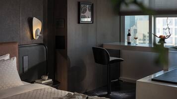 A modern hotel room with a bed, a desk, a chair, a window with a city view, and a wall-mounted light fixture.