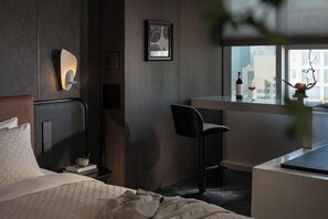 Moderni hotellihuone, jossa on sänky, työpöytä, tuoli, ikkuna, josta on näkymä kaupunkiin, ja seinään kiinnitetty valaisin.