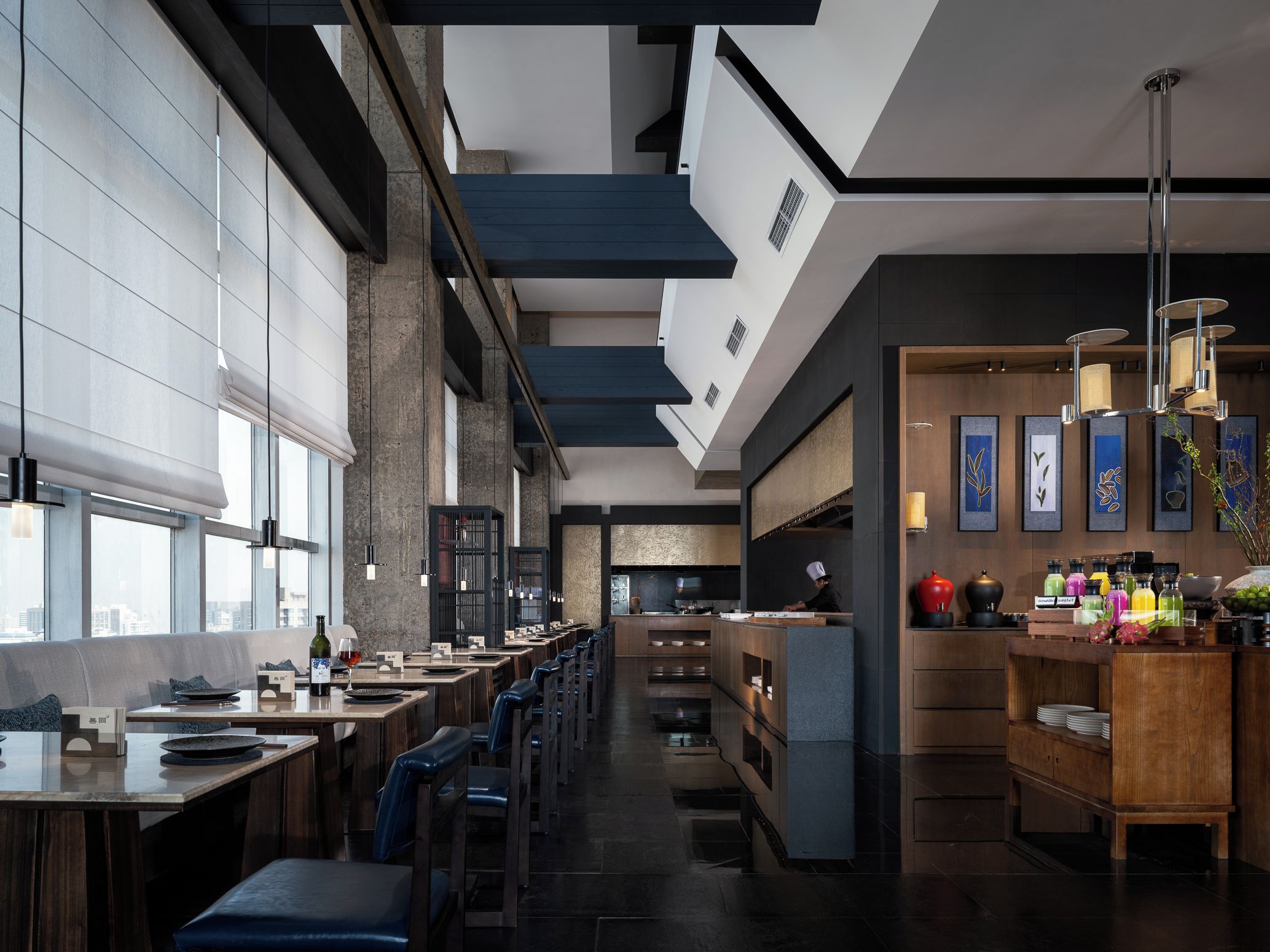 A modern restaurant interior with high ceilings, long communal tables, and a bar area.