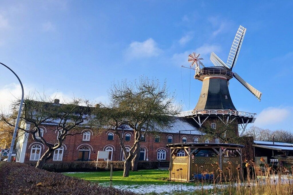 Un moulin à vent traditionnel, avec un bâtiment en briques en arrière-plan, une structure en bois au premier plan et un ciel bleu dégagé.