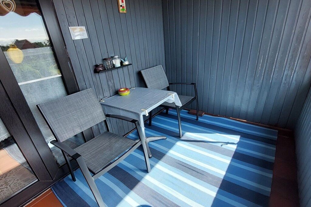 A small, enclosed outdoor seating area with a table and two chairs, a blue-striped rug, and a shelf with jars.