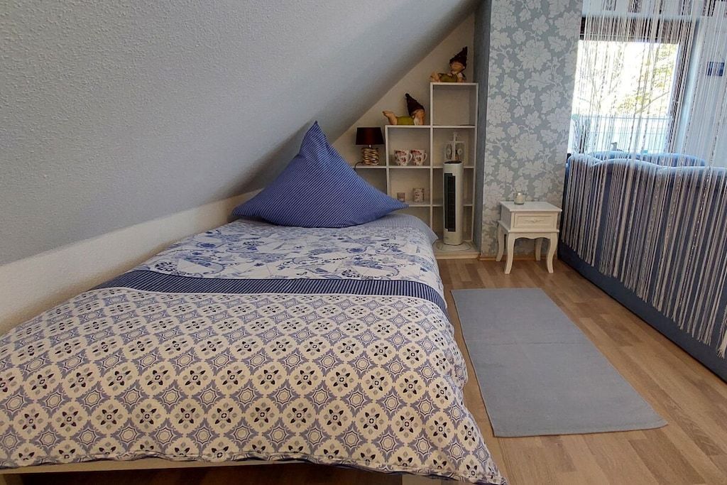 Een slaapkamer met een bed, een klein nachtkastje, een plank met decoratieve objecten en een raam met gordijnen.
