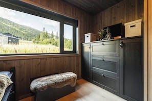 Interior de una cabaña con un banco de madera, un mueble con cajones, una ventana que ofrece vistas a un bosque y un edificio, y una pequeña estatua sobre el mueble.