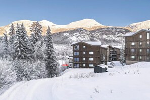 Een meerlaags houten gebouw met een skilift op de achtergrond, omgeven door besneeuwde bomen en bergen.