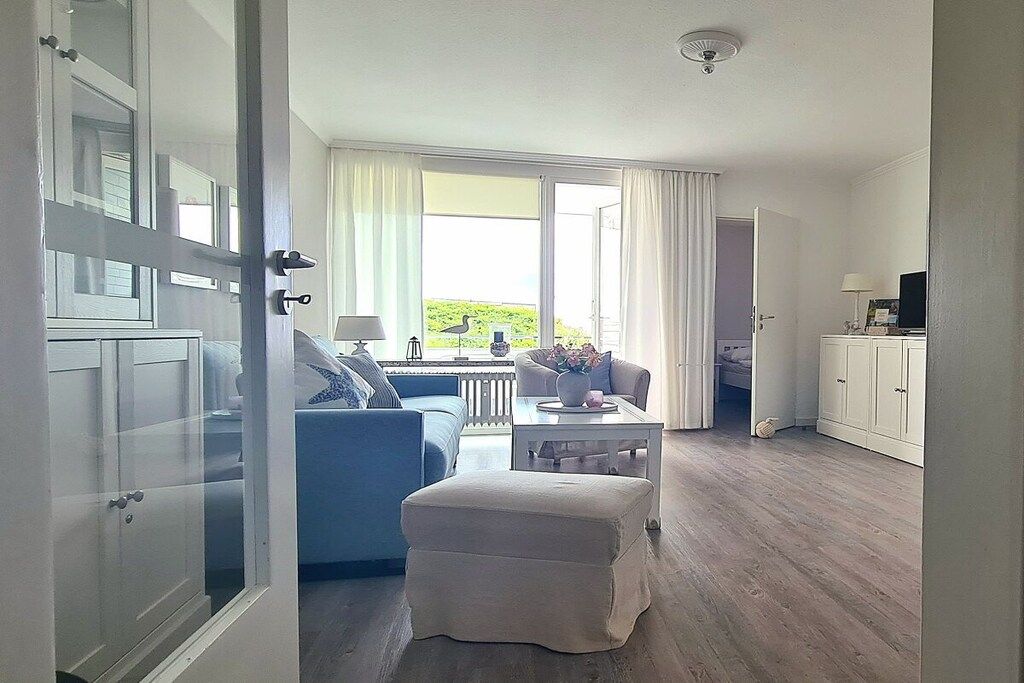 A room with a blue sofa, a white ottoman, a dining table, and a view through a glass door to the outside.