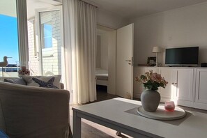 A room with a sofa, a table with a vase and candle, a television, and a door leading to another room. - Holiday apartment in Sylt with 2 bedrooms (Westerland\/Sylt)