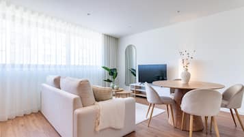 Un saló modern amb un sofà, una taula de dinar amb cadires, un televisió de pantalla plana i una finestra gran amb cortines primes.