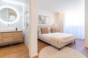 Quarto moderno com uma cama grande, um espelho redondo, um armário de madeira e uma janela ampla com cortinas leves.