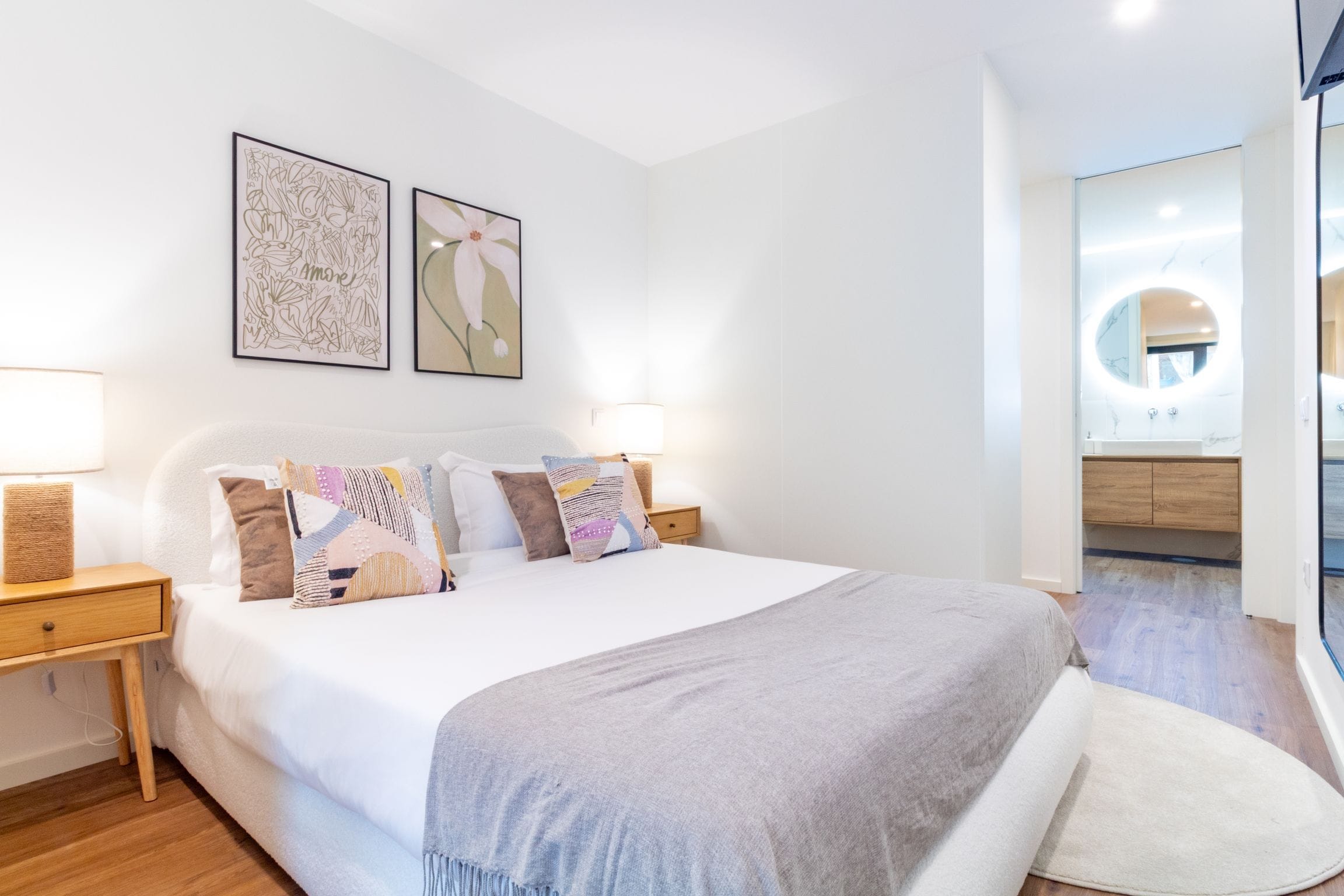 Une chambre à coucher moderne avec un grand lit, deux tables de chevet et une commode. Deux œuvres d’art encadrées ornent le mur, ainsi qu’un miroir rond. On aperçoit également une salle de bain avec un miroir rond.