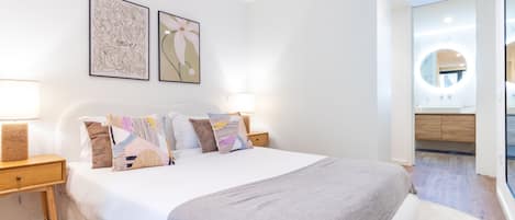 Une chambre à coucher moderne avec un grand lit, deux tables de chevet et une commode. Deux œuvres d’art encadrées ornent le mur, ainsi qu’un miroir rond. On aperçoit également une salle de bain avec un miroir rond.