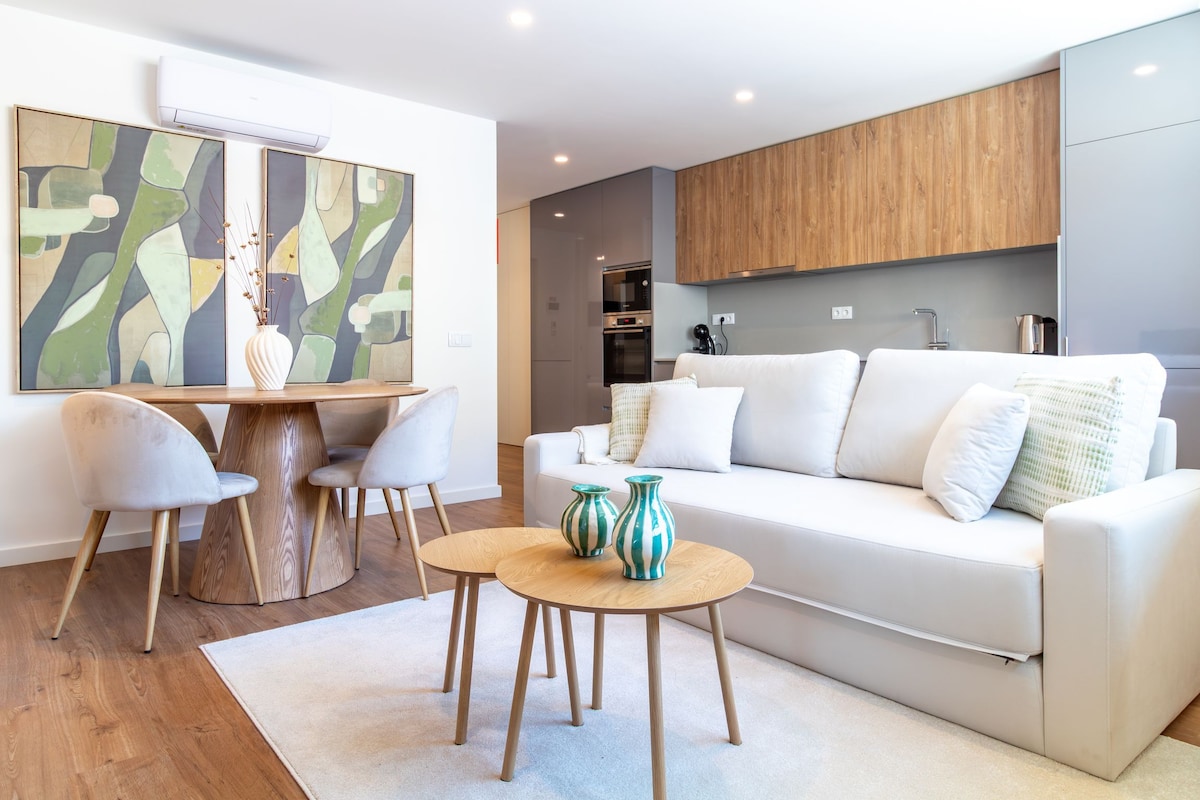 A modern living room with a white sofa, a round wooden coffee table, and two chairs. There is a painting on the wall and a kitchen area in the background.