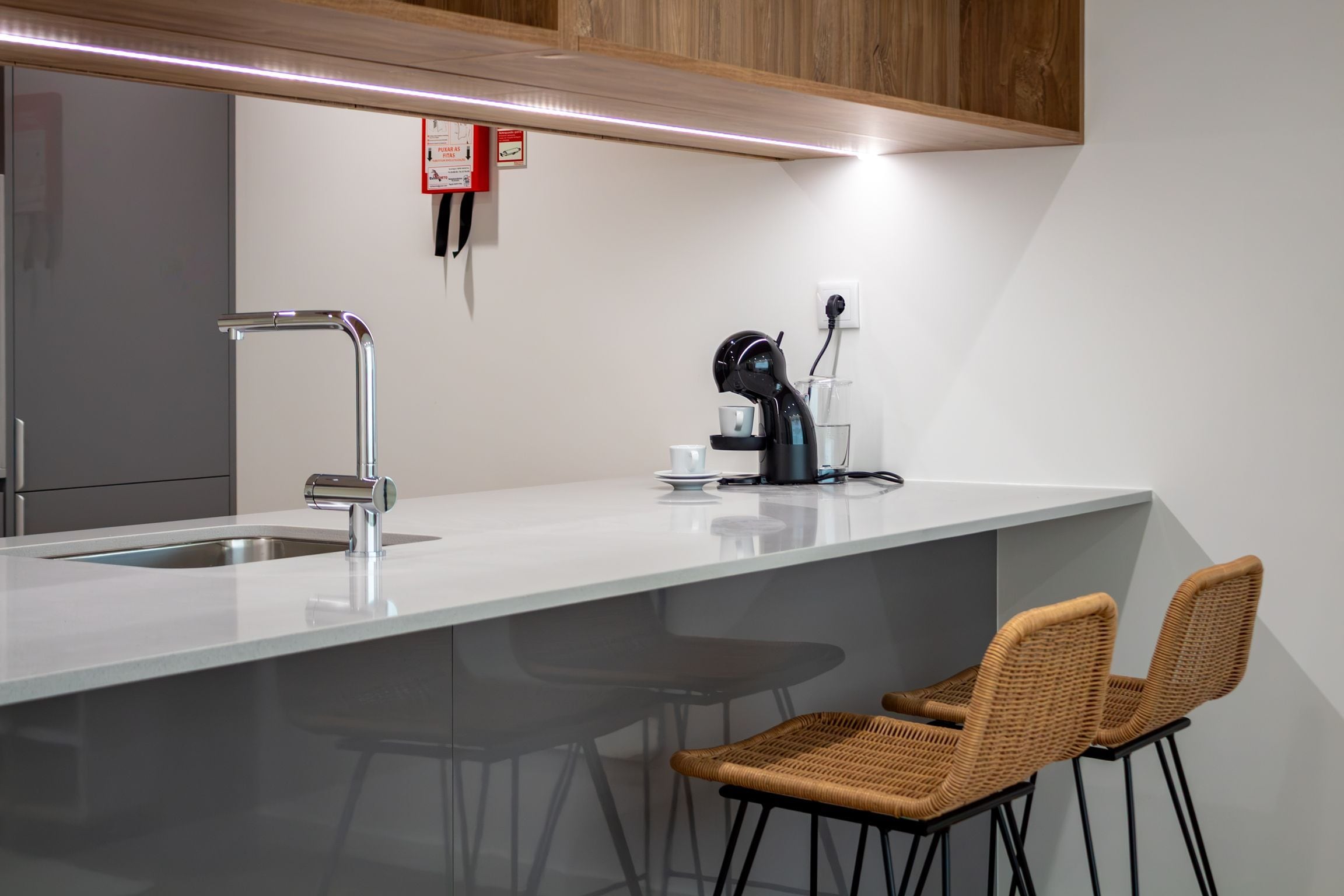 Una cocina moderna con encimera blanca, pileta de acero inoxidable y una máquina de café. Hay dos sillas de mimbre y una mesita con una taza y un platito.