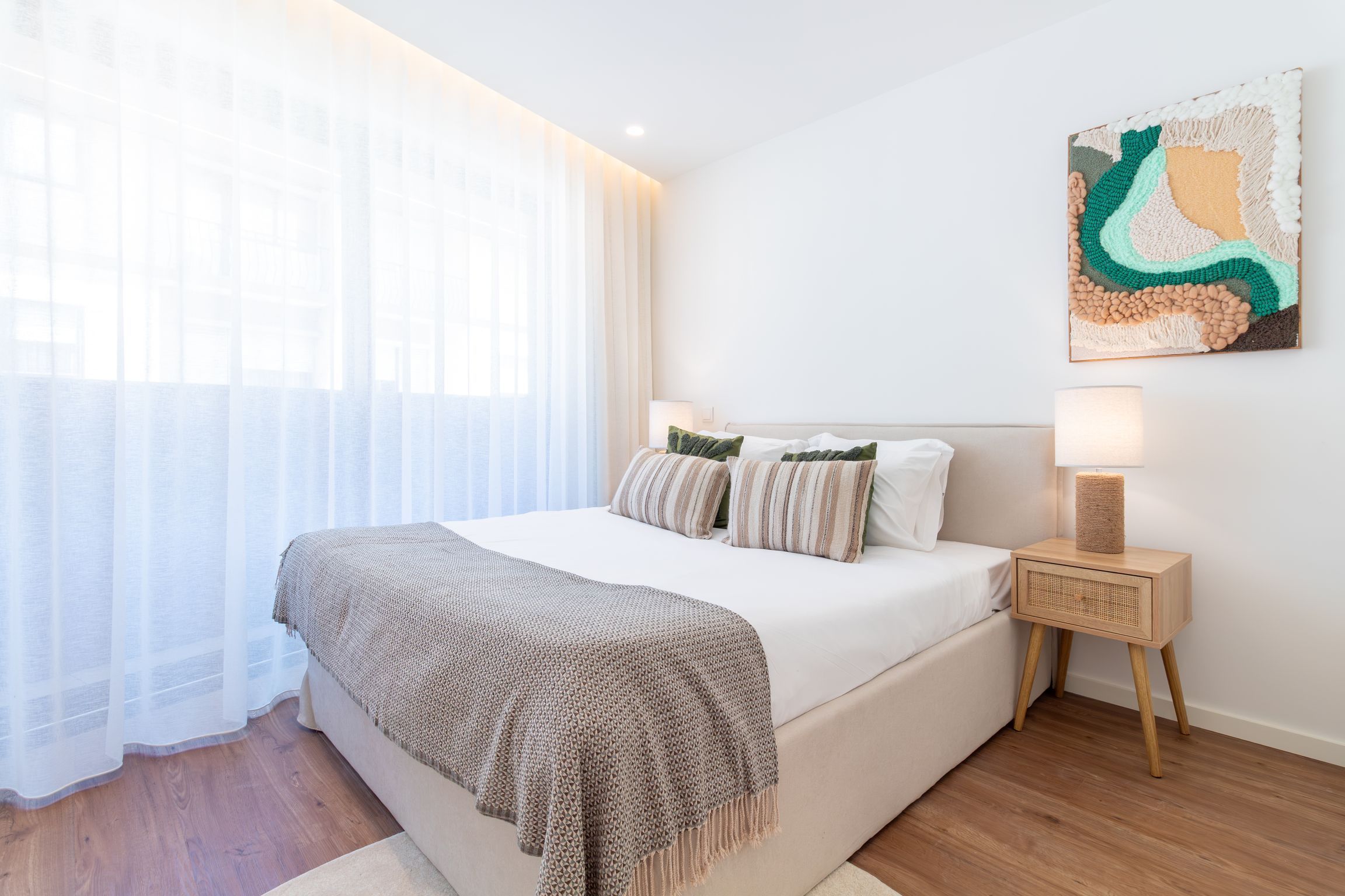 Een slaapkamer met een bed, nachtkastje, lamp en een abstract kunstwerk aan de muur.
