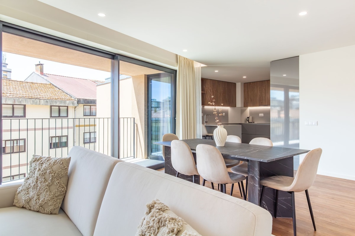 A modern apartment with a dining area, kitchen, and a large window overlooking a cityscape.